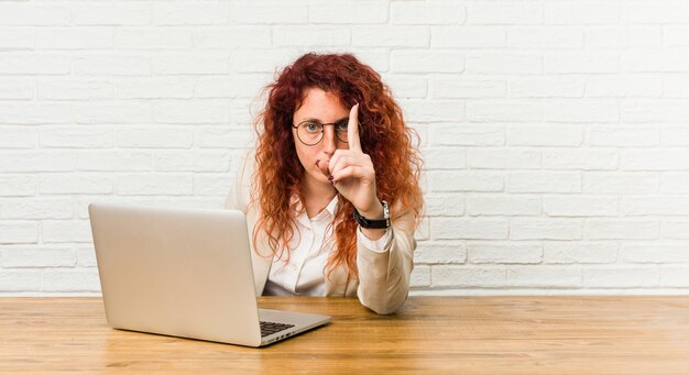 Donna riccia della giovane testarossa che lavora con il suo computer portatile che mostra numero uno con il dito.