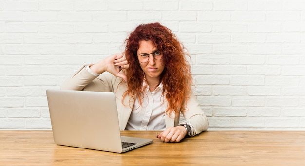 Donna riccia della giovane testarossa che lavora con il suo computer portatile che mostra un gesto di avversione, pollici giù