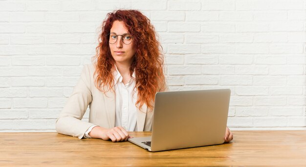 Donna riccia della giovane testarossa che lavora con il suo computer portatile felice, sorridente e allegro.