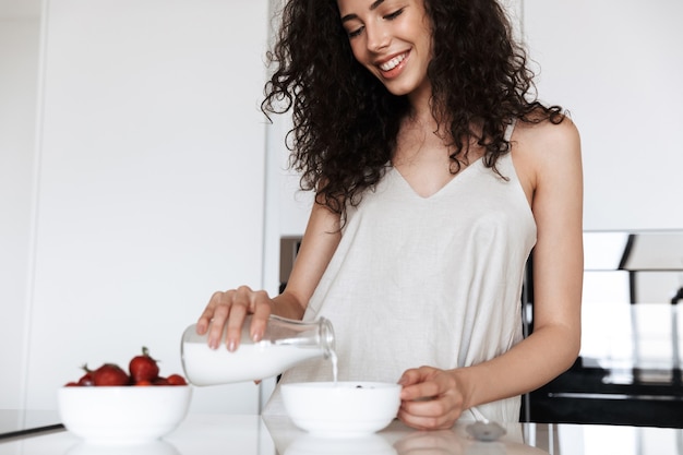 donna riccia felice che indossa abiti di seta per il tempo libero sorridente e versando il latte nel piatto in cucina interna, durante una sana colazione