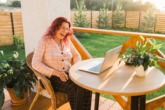 Donna ridente che indossa un pigiama durante una videochiamata sul laptop durante la pausa pranzo seduta in terrazza alle h