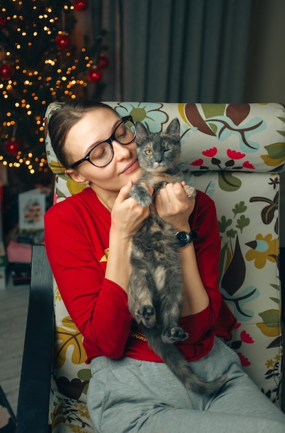 Donna rilassante in vetro che tiene un gatto vicino all'albero di Natale in una casa accogliente.