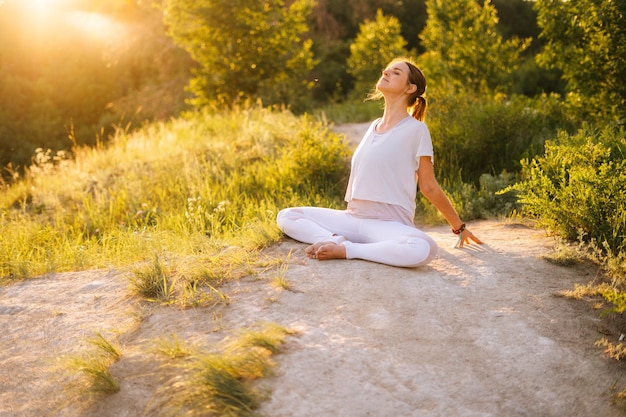 Donna rilassata seduta nella posa del loto e facendo piegare la schiena fuori nel parco sera sullo sfondo della luce del sole La pratica dello yoga femminile si esercita da sola all'aperto all'erba verde durante il tramonto