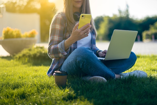 Donna ritagliata che manda un SMS allo smartphone seduto sull'erba fuori nel parco ragazza felice e sorridente che lavora