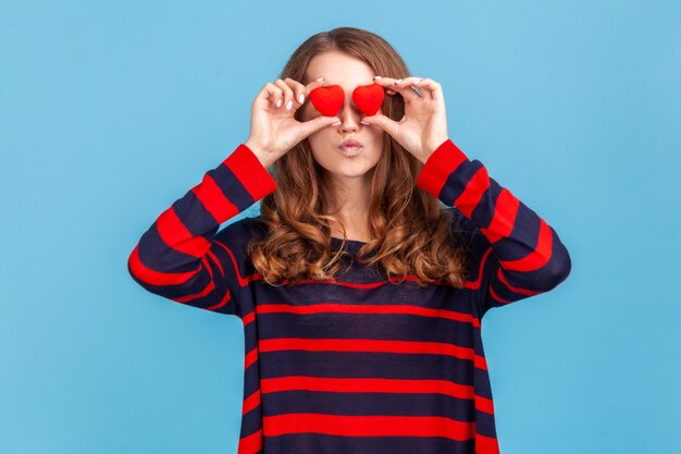 Donna romantica che indossa un maglione in stile casual a righe in piedi con le labbra imbronciate che inviano baci d'aria che coprono gli occhi con piccoli simboli d'amore del cuore rosso Colpo in studio al coperto isolato su sfondo blu