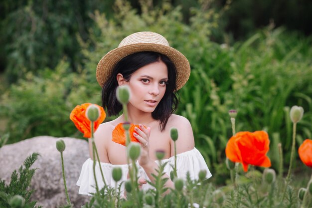 Donna romantica del brunette in cappello di paglia con il fiore a disposizione che si siede nel campo stupefacente del papavero.