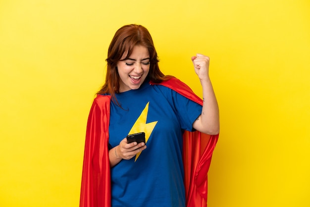 Donna rossa del super eroe isolata su fondo giallo con il telefono nella posizione di vittoria