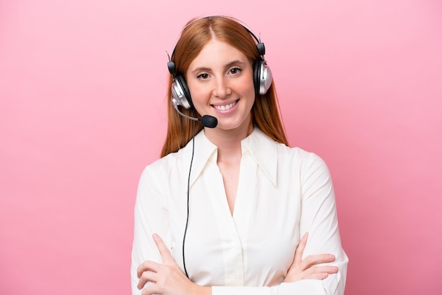 Donna rossa di telemarketing che lavora con un auricolare isolato su sfondo rosa mantenendo le braccia incrociate in posizione frontale