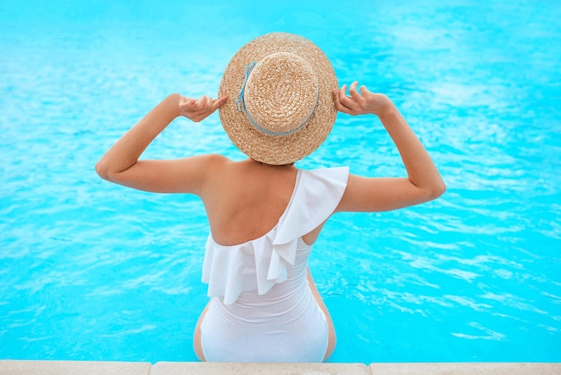 donna rossa è tornata in costume da bagno bianco seduta a bordo piscina con il cappello di paglia