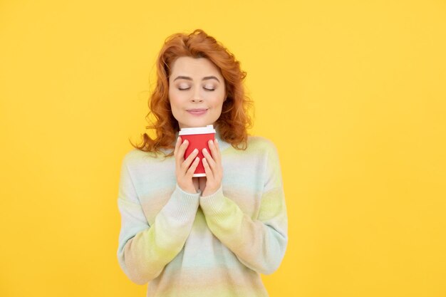 Donna rossa felice che sente l'odore del caffè da asporto dalla tazza su sfondo giallo buongiorno