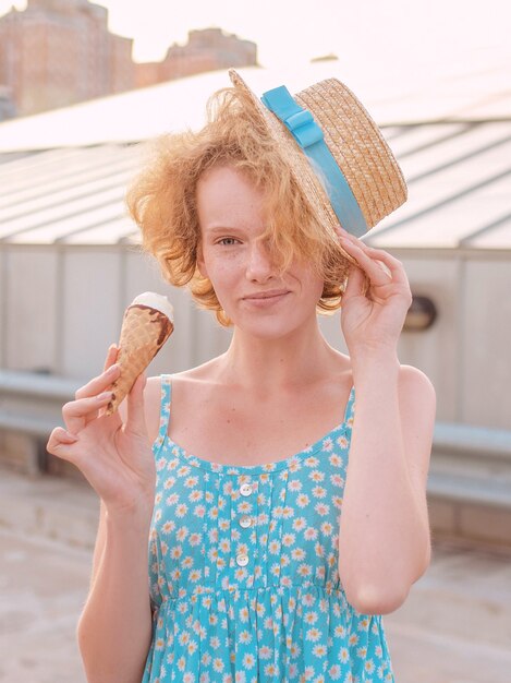 donna rossa riccia con cappello di paglia prendisole blu che mangia il gelato sul tetto del grattacielo skyroof