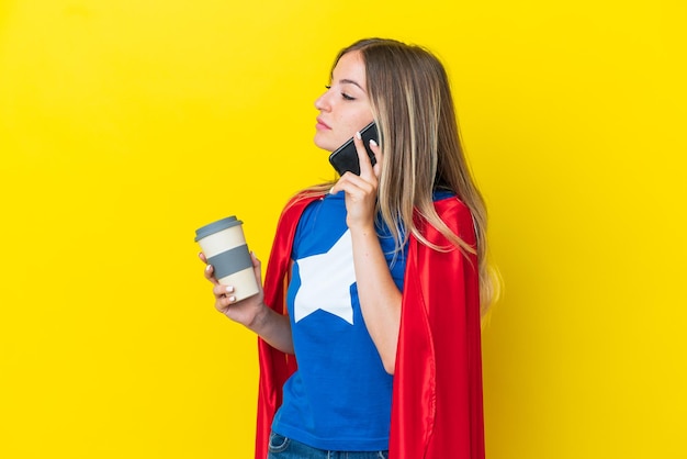 Donna rumena del supereroe isolata su fondo giallo che tiene il caffè da portare via e un cellulare