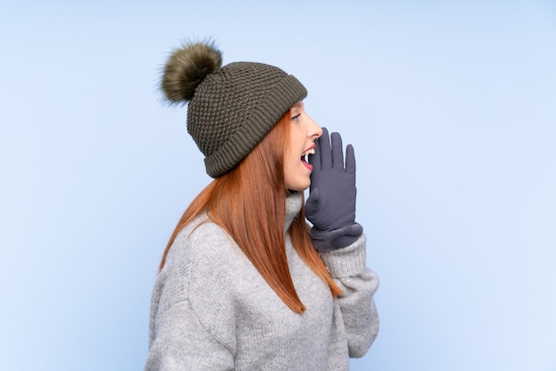 Donna russa della giovane testarossa con il cappello di inverno sopra la parete blu isolata che grida con la bocca spalancata