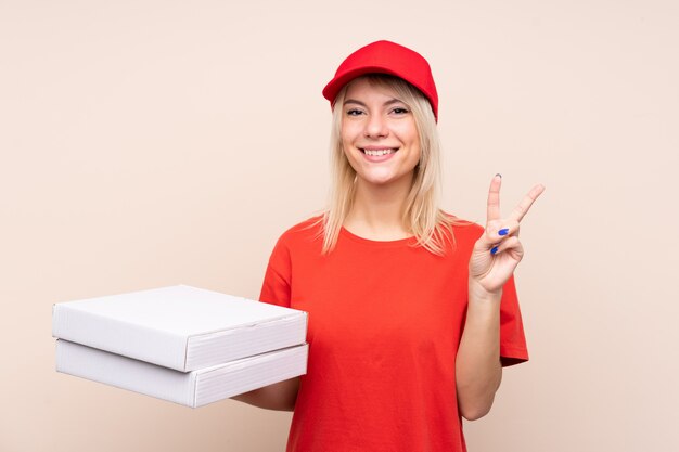 Donna russa di consegna della pizza che tiene una pizza sopra la parete isolata che sorride e che mostra il segno di vittoria