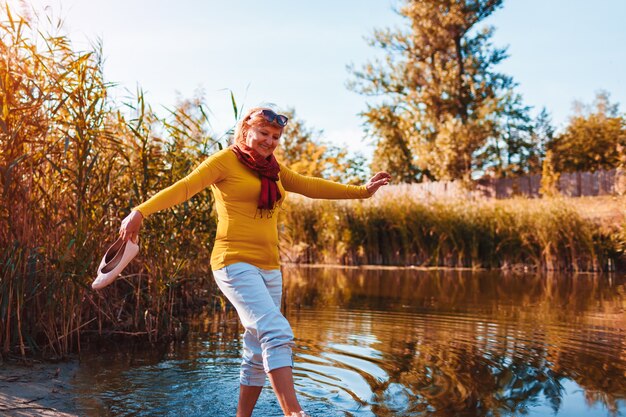 Donna scalza di mezza età che cammina sulla riva del fiume il giorno d'autunno. Signora anziana che si diverte nella foresta godendosi la natura