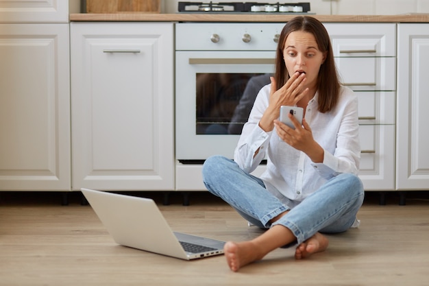 Donna scioccata che indossa camicia bianca e jeans, seduta sul pavimento in cucina vicino al notebook, tenendo in mano lo smartphone, guardando il display del dispositivo con espressione sorpresa, coprendo la bocca con il palmo.