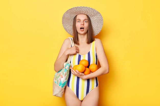 Donna scioccata che indossa il costume da bagno isolato su sfondo giallo che tiene le arance sorprendendosi del prezzo elevato per la frutta guardando la fotocamera con la bocca aperta