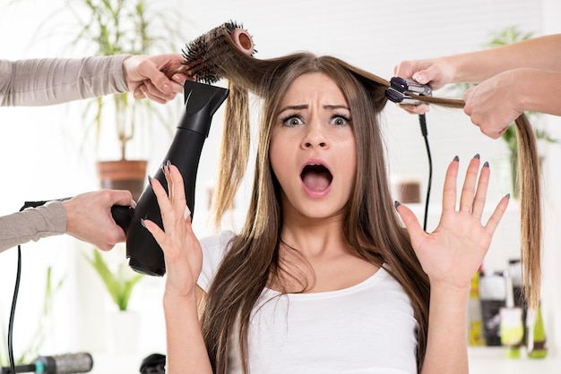 Donna scioccata che raddrizza lunghi capelli castani con asciugacapelli e spazzola rotonda e ferri da stiro dal parrucchiere.