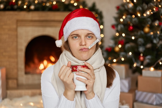 Donna sconvolta che indossa cappello, sciarpa e maglione di Babbo Natale, misurando la temperatura e bevendo tè caldo, tenendo la tazza con due mani, seduto vicino al camino e albero di Natale.