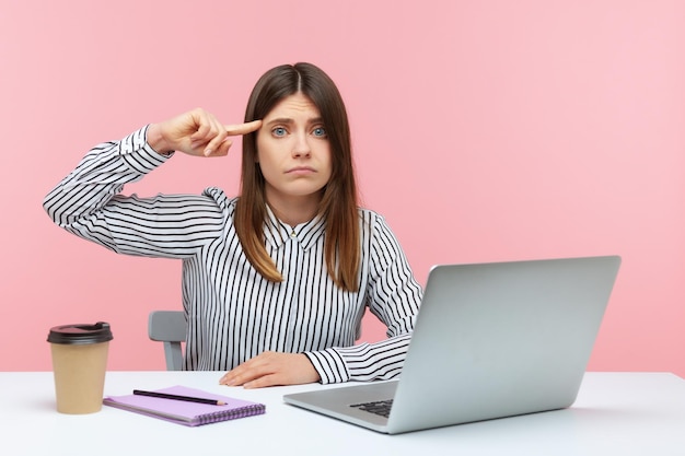 Donna sconvolta che mostra un gesto stupido con un dito che guarda la fotocamera con un'espressione stanca dispiaciuta, seduta sul posto di lavoro con il laptop. Studio indoor girato isolato su sfondo rosa