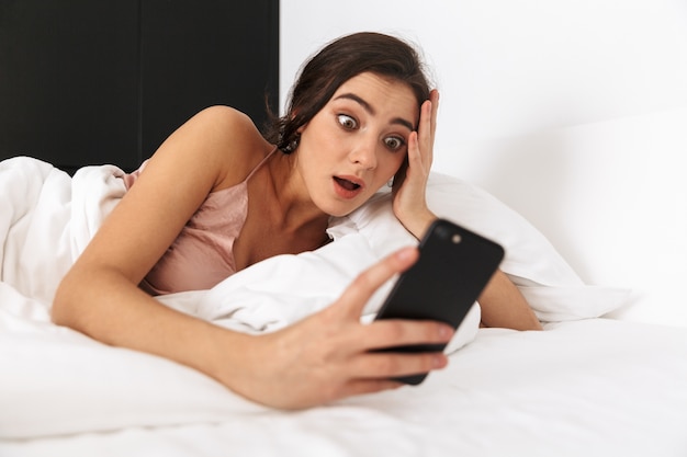 donna sconvolta guardando il telefono cellulare, mentre dorme nel letto su lino bianco