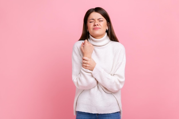 Donna sconvolta in piedi con una smorfia di dolore che massaggia il polso dolorante che soffre di lesioni alla mano o distorsione che indossa un maglione bianco in stile casual Colpo in studio al coperto isolato su sfondo rosa