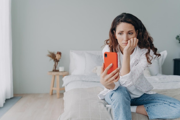 Donna sconvolta perplessa che legge cattive notizie sui media nei social network che tengono lo smartphone seduto in camera da letto