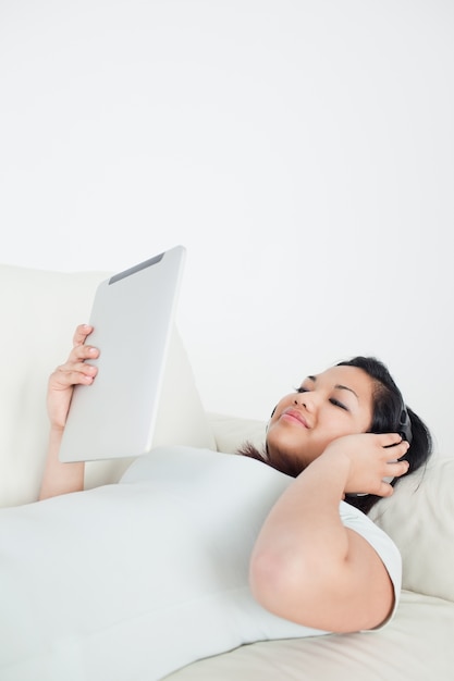 Donna sdraiata su un divano con le cuffie e in possesso di un tablet tattile