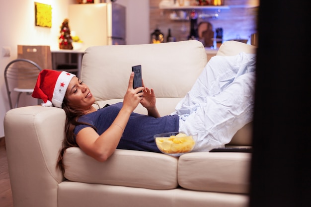 Donna sdraiata sul divano che naviga sui social media utilizzando lo smartphone durante il periodo natalizio
