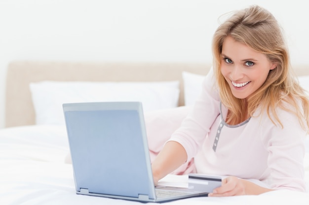 Donna sdraiata sul letto, con un portatile in mano e guardando avanti e sorridente