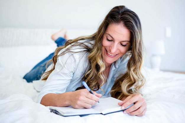 donna sdraiata sul suo letto, sorridendo e scrivendo in un libro con una penna
