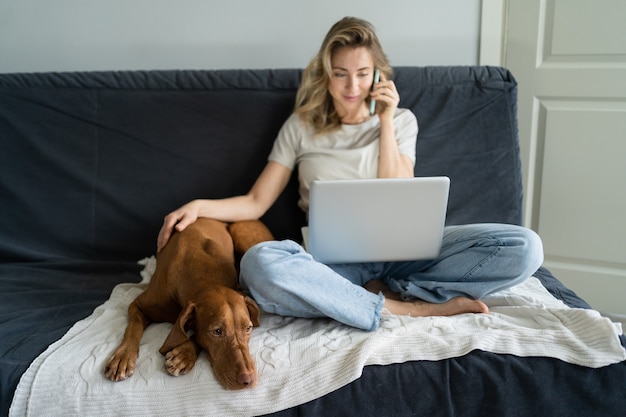 Donna seduta al divano con il cane, parlando al cellulare, lavorando in remoto sul computer portatile a casa