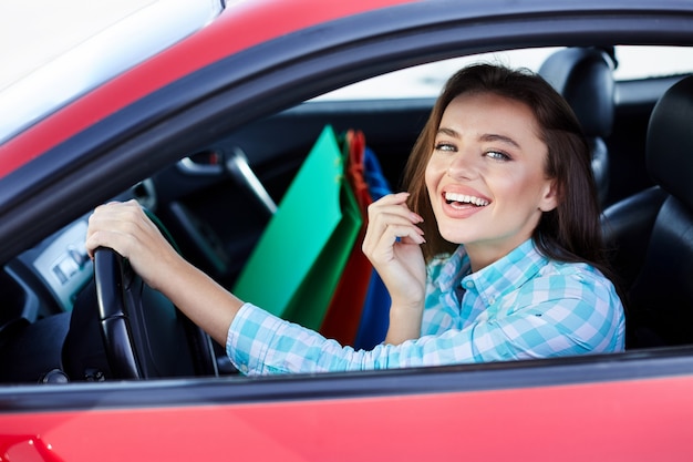 Donna seduta all'interno dell'auto rossa, autista felice. Donna che guarda la fotocamera e sorridente. Testa e spalle della donna felice che tiene il timone, tornando a fare shopping