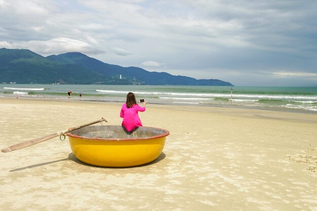 Donna seduta con il suo telefono cellulare sulla barca da pesca rotonda impermeabile di bambù presso la China Beach a Danang in Vietnam. Si chiama anche Spiaggia di Non Nuoc. Sullo sfondo il Mar Cinese Meridionale.