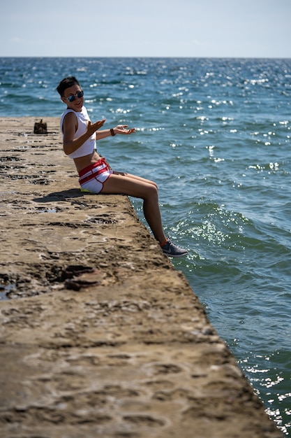 Donna seduta da sola sul molo in pantaloncini corti e canottiera con scarpe. Vista posteriore. Mare d'estate