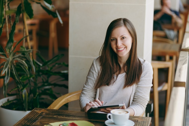 Donna seduta da sola vicino alla grande finestra nella caffetteria al tavolo con una tazza di cappuccino, torta, rilassante nel ristorante durante il tempo libero. Donna che lavora al computer tablet pc resto nella caffetteria. Concetto di stile di vita.