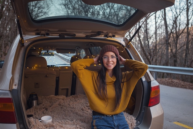 Donna seduta nel retro del bagagliaio dell'auto. Prepararsi per partire. Giovane donna che ride seduta nel bagagliaio aperto della sua auto. Viaggio on the road autunnale. Viaggio hipster rospo, fuga autunnale