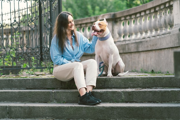 Donna seduta su una scala di pietra e accarezzando il cane felice dell'american pitbull terrier guardando il proprietario