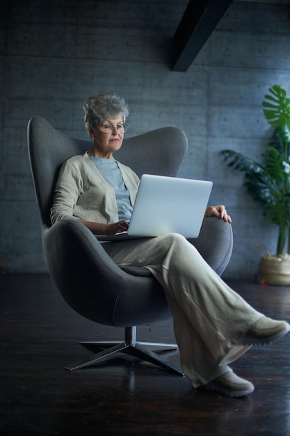 Donna seduta su una sedia moderna vicino alla finestra in una stanza luminosa e accogliente a casa, sta lavorando su un laptop in...