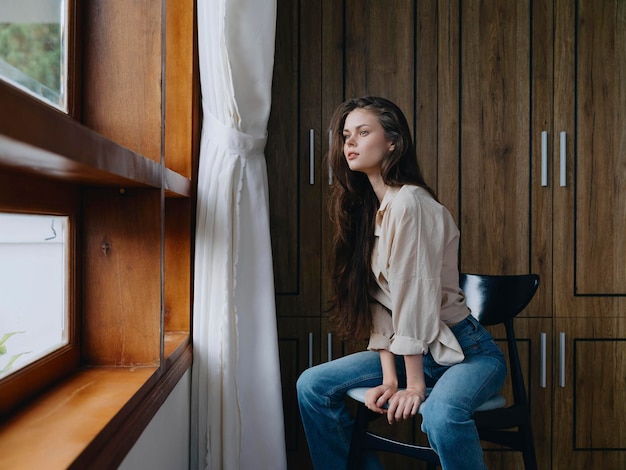 Donna seduta su una sedia nell'interno della casa vicino alla finestra conforto e comodità stile di vita relax nel fine settimana sguardo pensieroso di una bella ragazza