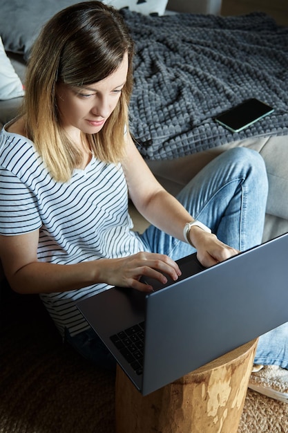 Donna seduta sul divano in soggiorno e utilizzando il computer portatile femminile freelance che lavora a casa ufficio remoto