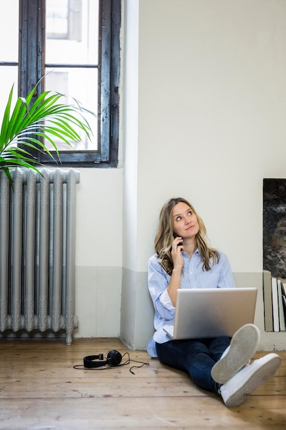 Donna seduta sul pavimento a casa tramite telefono cellulare e laptop