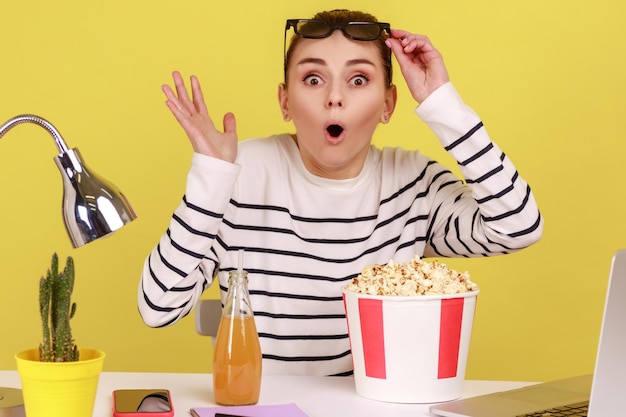 Donna seduta sul posto di lavoro con popcorn e bevande alzando i bicchieri e guardando un momento scioccante