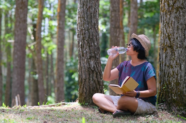 Donna seduta sull'erba sotto un pino che beve una bottiglia di acqua minerale mentre legge un libro