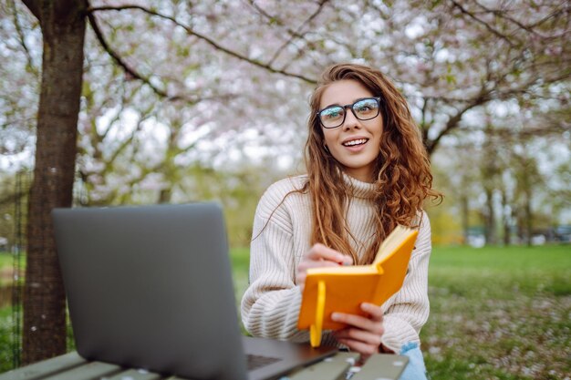 Donna seduta sull'erba verde vicino agli alberi in fiore con il portatile scrive nel blocco appunti concetto di pianificazione