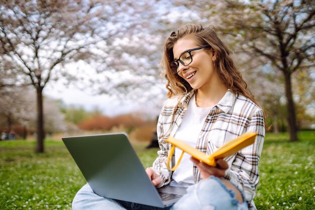 Donna seduta sull'erba verde vicino agli alberi in fiore con il portatile scrive nel blocco appunti concetto di pianificazione