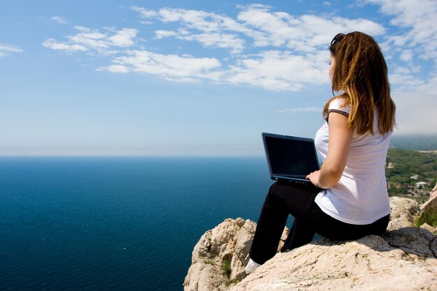 Donna seduta sulla roccia con il computer portatile vicino al mare