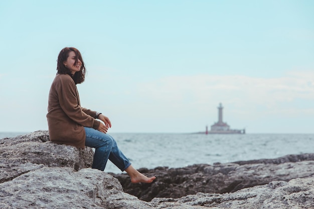 Donna seduta sulla spiaggia rocciosa del mare in jeans bagnati faro su sfondo tempo ventoso vacanze estive concetto spensierato