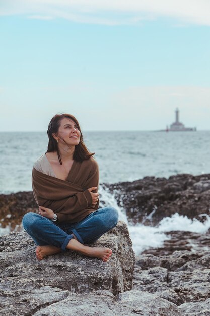 Donna seduta sulla spiaggia rocciosa del mare in jeans bagnati faro sullo sfondo