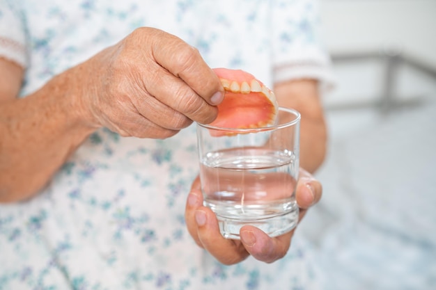 Donna senior asiatica che tiene e lava la protesi dentaria in un bicchiere con acqua detergente per una buona masticazione